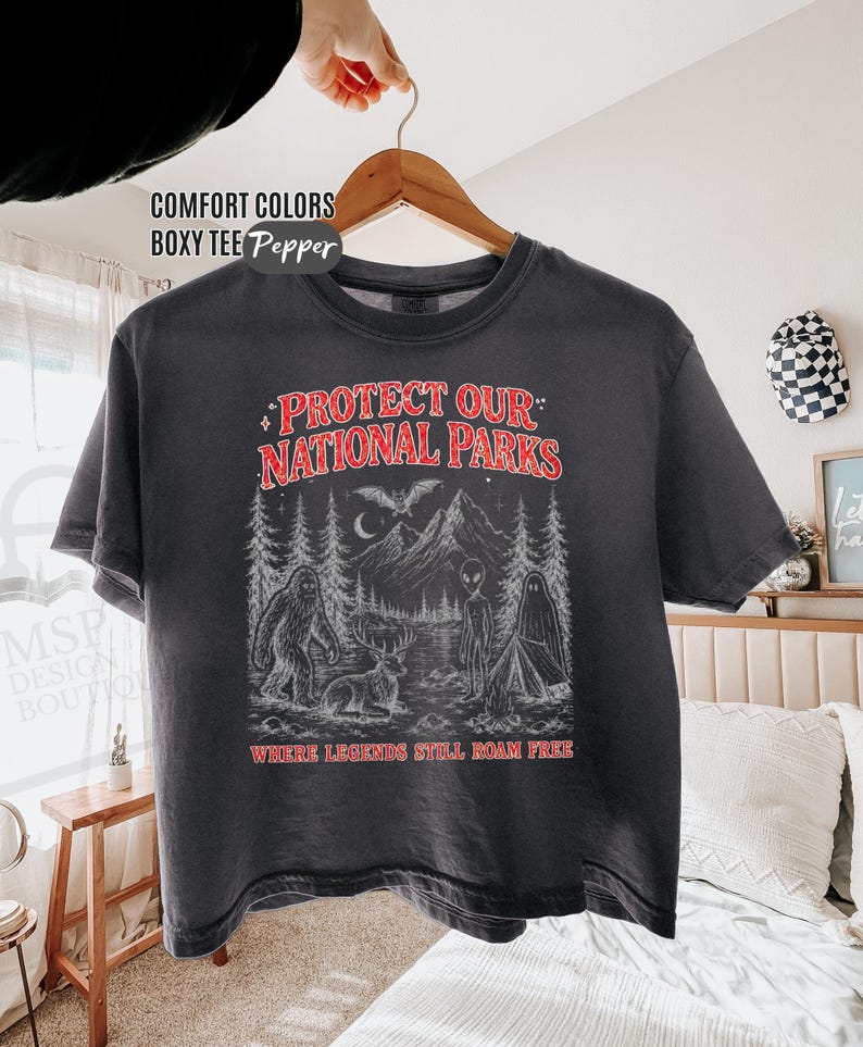 May include: A charcoal gray boxy t-shirt featuring a graphic print with the text "PROTECT OUR NATIONAL PARKS" and "WHERE LEGENDS STILL ROAM FREE" in red. The illustration depicts a forest, mountains, a bat, a deer, a ghost, and an alien.
