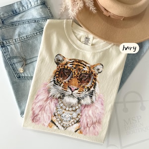 May include: An ivory t-shirt featuring a graphic illustration of a tiger wearing leopard print sunglasses, a pink faux fur stole, and layered crystal necklaces. The shirt includes a Comfort Colors label and the text Ivory printed near the top.