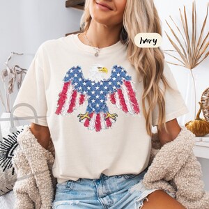 May include: An ivory short-sleeve t-shirt featuring a graphic of a bald eagle with wings spread. The eagle is designed with a distressed American flag pattern in red, white, and blue. The text "IVORY" is printed in a small circle on the upper right.