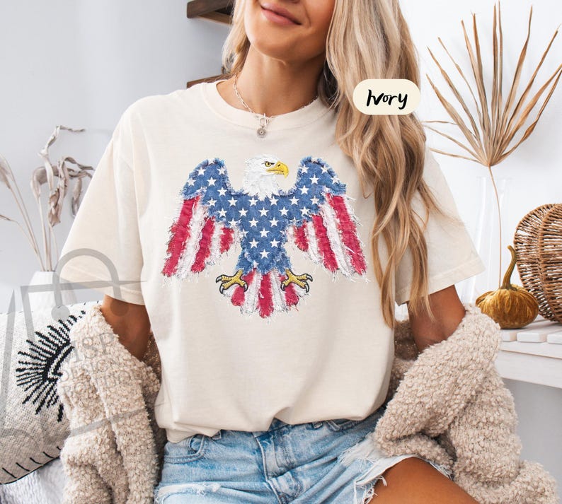 May include: An ivory short-sleeve t-shirt featuring a graphic of a bald eagle with wings spread. The eagle is designed with a distressed American flag pattern in red, white, and blue. The text "IVORY" is printed in a small circle on the upper right.