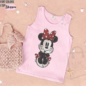 May include: A light pink tank top featuring a graphic print of Minnie Mouse with a red and white polka dot bow. The text COMFORT COLORS TANK TOP Blossom is printed in the upper left corner. A light pink woven handbag is positioned to the left.