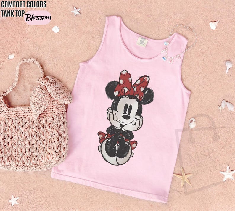 May include: A light pink tank top featuring a graphic print of Minnie Mouse with a red and white polka dot bow. The text COMFORT COLORS TANK TOP Blossom is printed in the upper left corner. A light pink woven handbag is positioned to the left.