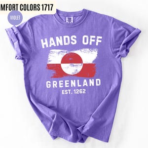 May include: A violet t-shirt with the words "HANDS OFF GREENLAND" in white, along with a Greenland flag design. The text "EST. 1262" is below the flag. The shirt is labeled "COMFORT COLORS 1717" and "VIOLET".