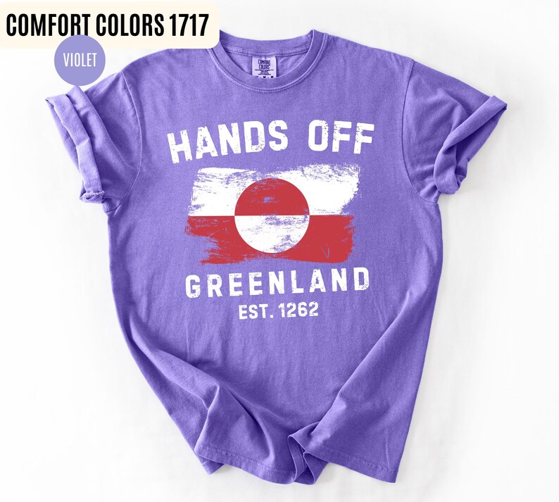 May include: A violet t-shirt with the words "HANDS OFF GREENLAND" in white, along with a Greenland flag design. The text "EST. 1262" is below the flag. The shirt is labeled "COMFORT COLORS 1717" and "VIOLET".