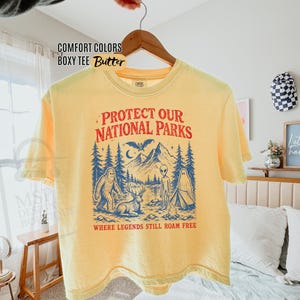 May include: A butter yellow boxy t-shirt featuring a graphic print in red and blue. The design includes mountains, trees, a bat, a bigfoot, a deer, an alien, and a ghost by a campfire with the text "PROTECT OUR NATIONAL PARKS WHERE LEGENDS STILL ROAM FREE."