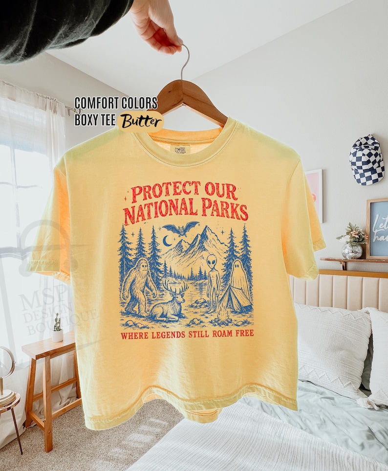 May include: A butter yellow boxy t-shirt featuring a graphic print in red and blue. The design includes mountains, trees, a bat, a bigfoot, a deer, an alien, and a ghost by a campfire with the text "PROTECT OUR NATIONAL PARKS WHERE LEGENDS STILL ROAM FREE."
