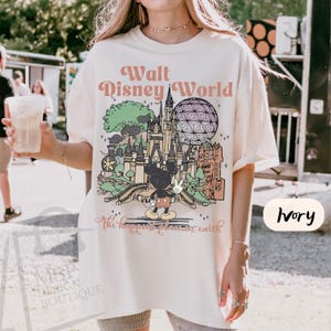 May include: Ivory-colored t-shirt featuring a colorful graphic of Walt Disney World landmarks, including Cinderella's castle and Spaceship Earth. The text "Walt Disney World" and "The happiest place on earth" are printed on the shirt. The shirt is a relaxed fit.