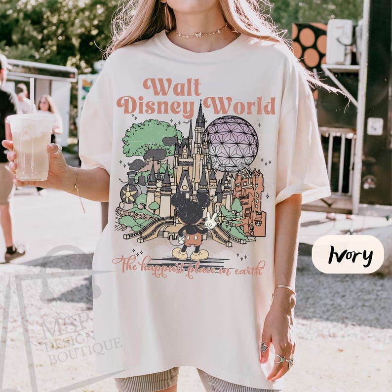 May include: Ivory-colored t-shirt featuring a colorful graphic of Walt Disney World landmarks, including Cinderella's castle and Spaceship Earth. The text "Walt Disney World" and "The happiest place on earth" are printed on the shirt. The shirt is a relaxed fit.