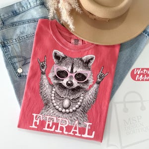 May include: A red t-shirt with a raccoon graphic wearing pink sunglasses and a pearl necklace, with the word "FERAL" in white. The shirt is on top of blue jeans, with a tan hat and a gold necklace.