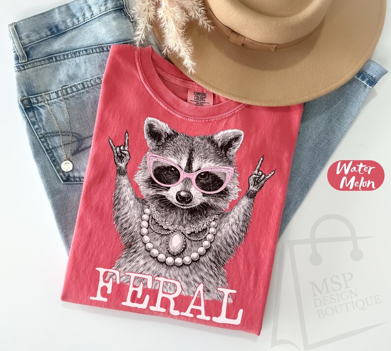 May include: A red t-shirt with a raccoon graphic wearing pink sunglasses and a pearl necklace, with the word "FERAL" in white. The shirt is on top of blue jeans, with a tan hat and a gold necklace.