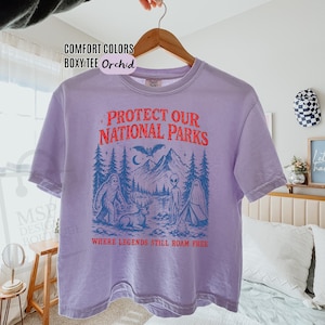 May include: A light purple boxy t-shirt featuring a graphic print with the text "PROTECT OUR NATIONAL PARKS" in red and "WHERE LEGENDS STILL ROAM FREE" in orange. The illustration depicts a forest, mountains, a bat, a yeti, a deer, an alien, and a ghost.