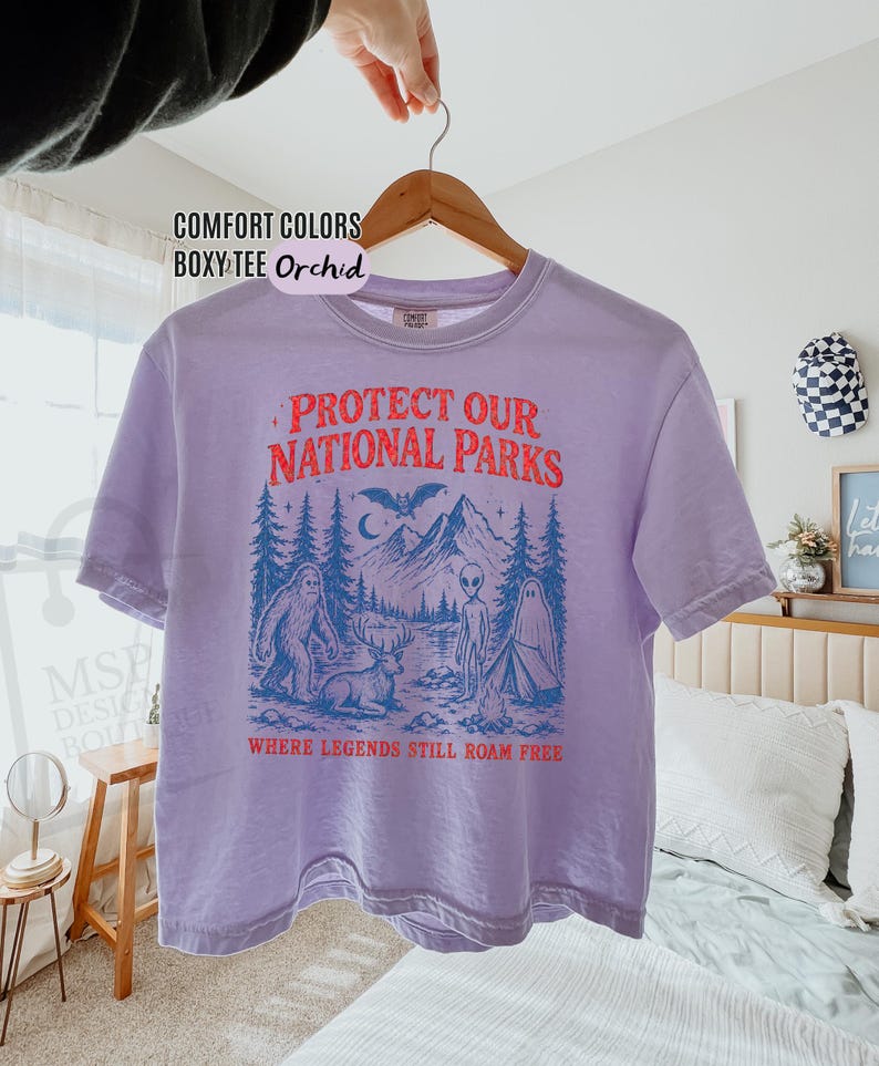 May include: A light purple boxy t-shirt featuring a graphic print with the text "PROTECT OUR NATIONAL PARKS" in red and "WHERE LEGENDS STILL ROAM FREE" in orange. The illustration depicts a forest, mountains, a bat, a yeti, a deer, an alien, and a ghost.