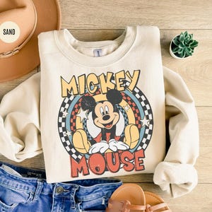Puede incluir: Sudadera color crema con un gr&aacute;fico retro de Mickey Mouse. El dise&ntilde;o incluye a Mickey en una pose sentada, rodeado de un arco&iacute;ris y estrellas, con el texto "Mickey Mouse". La sudadera se combina con pantalones cortos de mezclilla, un sombrero y sandalias.