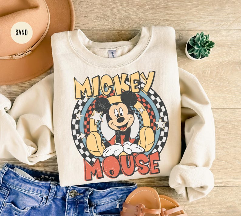 Puede incluir: Sudadera color crema con un gr&aacute;fico retro de Mickey Mouse. El dise&ntilde;o incluye a Mickey en una pose sentada, rodeado de un arco&iacute;ris y estrellas, con el texto "Mickey Mouse". La sudadera se combina con pantalones cortos de mezclilla, un sombrero y sandalias.