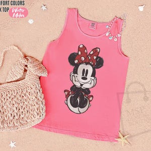 May include: A watermelon pink Comfort Colors tank top featuring a graphic print of Minnie Mouse with a red and white polka dot bow. The tank top has a round neckline and sleeveless design. Text on the garment reads Comfort Colors Tank Top.