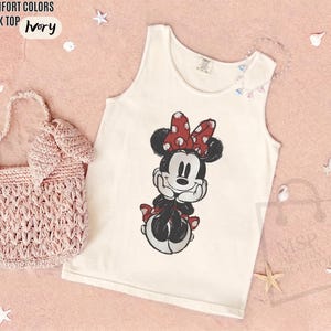 May include: An ivory tank top featuring a graphic print of Minnie Mouse with a red and white polka dot bow. The tank top is displayed alongside a light pink crocheted handbag. Text on the image reads COMFORT COLORS TANK TOP IVORY and MSP DESIGN BOUTIQUE.