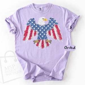 May include: A light purple short-sleeve t-shirt featuring a graphic of a bald eagle with wings and tail feathers patterned like the American flag. The eagle has a white head and yellow beak and talons. The word Orchid is printed on the right side.