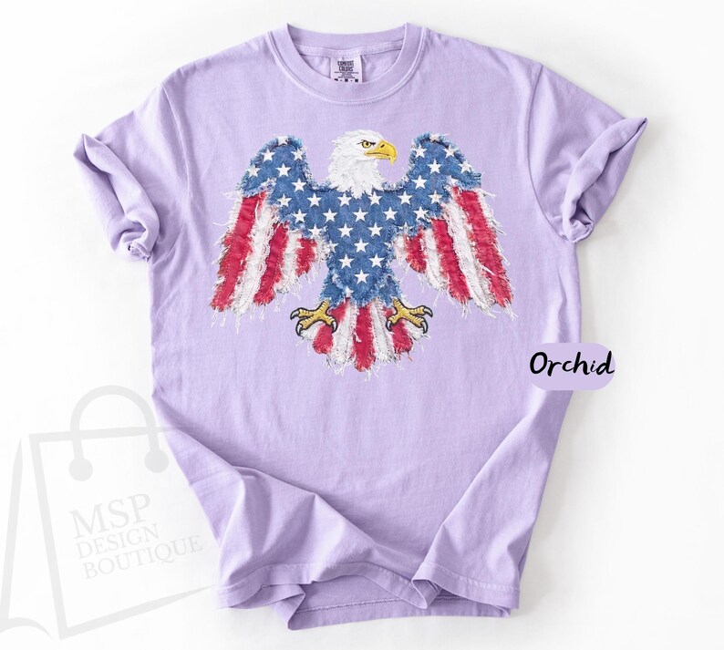May include: A light purple short-sleeve t-shirt featuring a graphic of a bald eagle with wings and tail feathers patterned like the American flag. The eagle has a white head and yellow beak and talons. The word Orchid is printed on the right side.