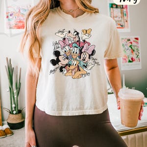 Puede incluir: Camiseta holgada de color marfil con un gr&aacute;fico colorido de personajes de Disney, incluyendo Mickey Mouse, Minnie Mouse, Donald Duck y Pluto. La camiseta tiene el texto "COMFORT COLORS - BOXY TEE" en la parte superior.