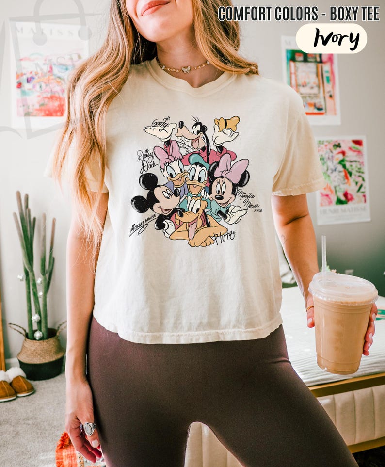 Puede incluir: Camiseta holgada de color marfil con un gr&aacute;fico colorido de personajes de Disney, incluyendo Mickey Mouse, Minnie Mouse, Donald Duck y Pluto. La camiseta tiene el texto "COMFORT COLORS - BOXY TEE" en la parte superior.