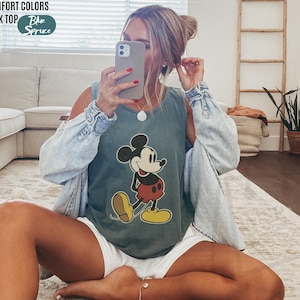 May include: A blue-green tank top featuring a classic Mickey Mouse graphic. The tank top is paired with a light blue denim jacket. The words "COMFORT COLORS TANK TOP" and "Blue Spruce" are printed on the top left.