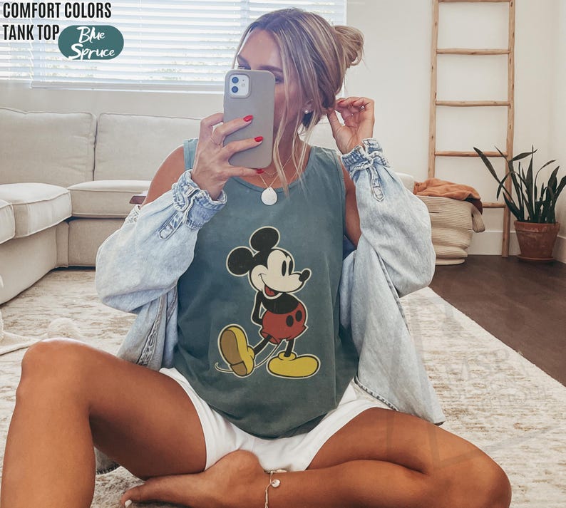 May include: A blue-green tank top featuring a classic Mickey Mouse graphic. The tank top is paired with a light blue denim jacket. The words "COMFORT COLORS TANK TOP" and "Blue Spruce" are printed on the top left.