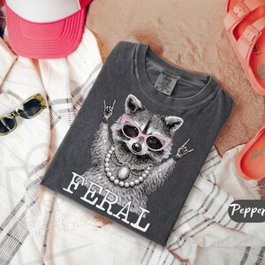 May include: Dark grey t-shirt featuring a raccoon wearing pink sunglasses and a pearl necklace, with the word "FERAL" in white. The raccoon is making a rock and roll hand gesture. The shirt is folded on a patterned fabric.