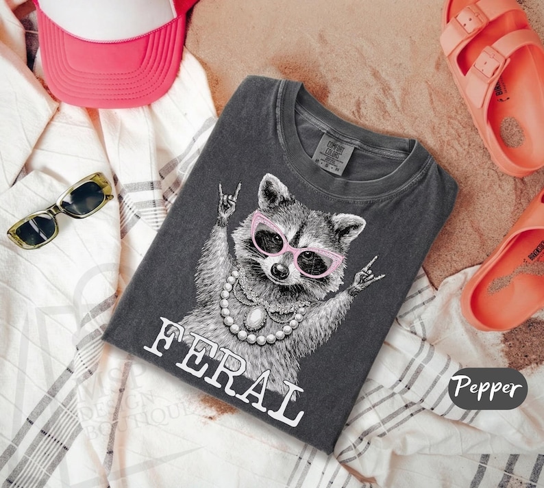 May include: Dark grey t-shirt featuring a raccoon wearing pink sunglasses and a pearl necklace, with the word "FERAL" in white. The raccoon is making a rock and roll hand gesture. The shirt is folded on a patterned fabric.