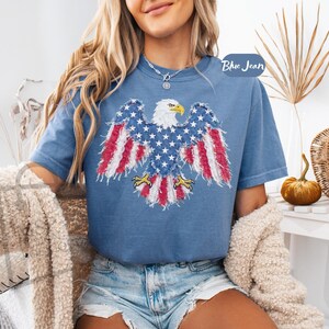 May include: A blue short-sleeved t-shirt featuring a graphic of a bald eagle with wings spread, designed with a distressed American flag pattern in red, white, and blue. The eagle has a yellow beak and talons. The text Blue Jean is visible.
