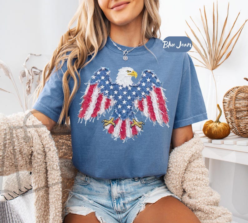 May include: A blue short-sleeved t-shirt featuring a graphic of a bald eagle with wings spread, designed with a distressed American flag pattern in red, white, and blue. The eagle has a yellow beak and talons. The text Blue Jean is visible.