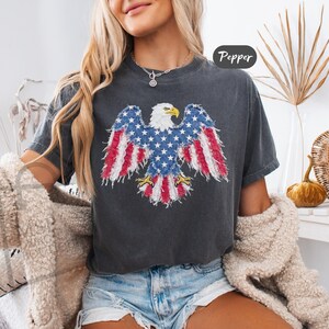 May include: A charcoal gray short-sleeve t-shirt featuring a graphic of a bald eagle with wings spread. The eagle's wings and tail are patterned with red and white stripes and white stars on a blue background. The text Pepper is visible in the corner.
