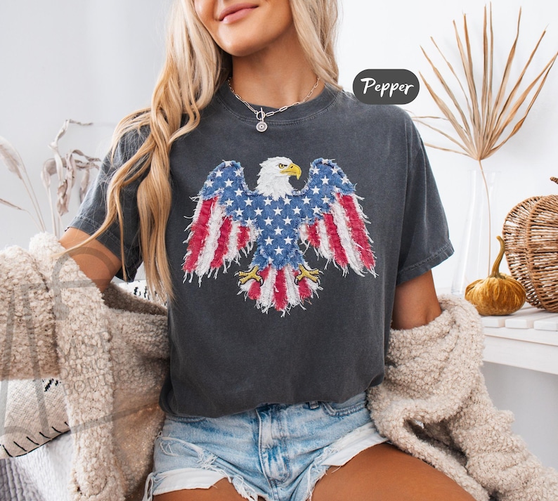 May include: A charcoal gray short-sleeve t-shirt featuring a graphic of a bald eagle with wings spread. The eagle's wings and tail are patterned with red and white stripes and white stars on a blue background. The text Pepper is visible in the corner.