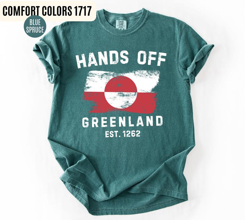 May include: Teal-colored t-shirt with the words "HANDS OFF GREENLAND" in white block letters. The shirt features a distressed Greenland flag design in red, white, and a red circle. The text "EST. 1262" is below the flag.
