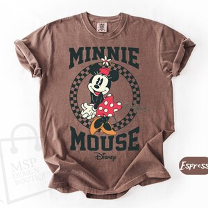 May include: Brown t-shirt featuring a graphic of Minnie Mouse with the text "MINNIE MOUSE" in dark green. Minnie is wearing a red and white polka dot dress. The shirt is a crew neck and has rolled sleeves.