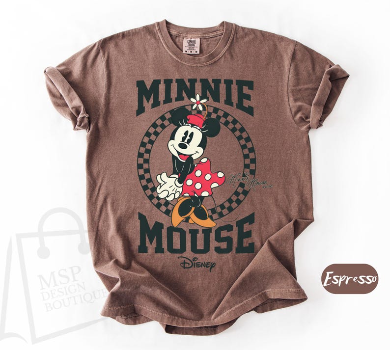 May include: Brown t-shirt featuring a graphic of Minnie Mouse with the text "MINNIE MOUSE" in dark green. Minnie is wearing a red and white polka dot dress. The shirt is a crew neck and has rolled sleeves.