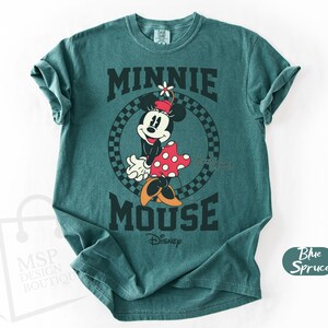 May include: Teal-colored t-shirt featuring a graphic of Minnie Mouse. The design includes Minnie in a red and white polka-dot dress, a flower on her head, and the words "MINNIE MOUSE" in bold, black letters. The Disney logo is also visible.