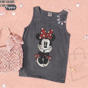 May include: A charcoal gray tank top featuring a graphic of Minnie Mouse with a red and white polka dot bow. The text "COMFORT COLORS TANK TOP" and "Pepper" appears in the upper left corner. A light pink woven handbag is positioned to the left.