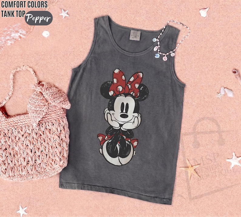 May include: A charcoal gray tank top featuring a graphic of Minnie Mouse with a red and white polka dot bow. The text "COMFORT COLORS TANK TOP" and "Pepper" appears in the upper left corner. A light pink woven handbag is positioned to the left.