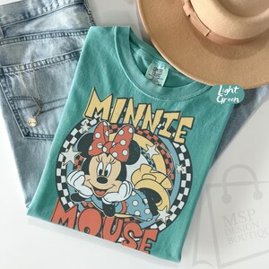 May include: A light green t-shirt featuring a graphic of Minnie Mouse with a red polka dot bow. The shirt is paired with light blue jeans and a tan fedora hat. A gold necklace is also visible.