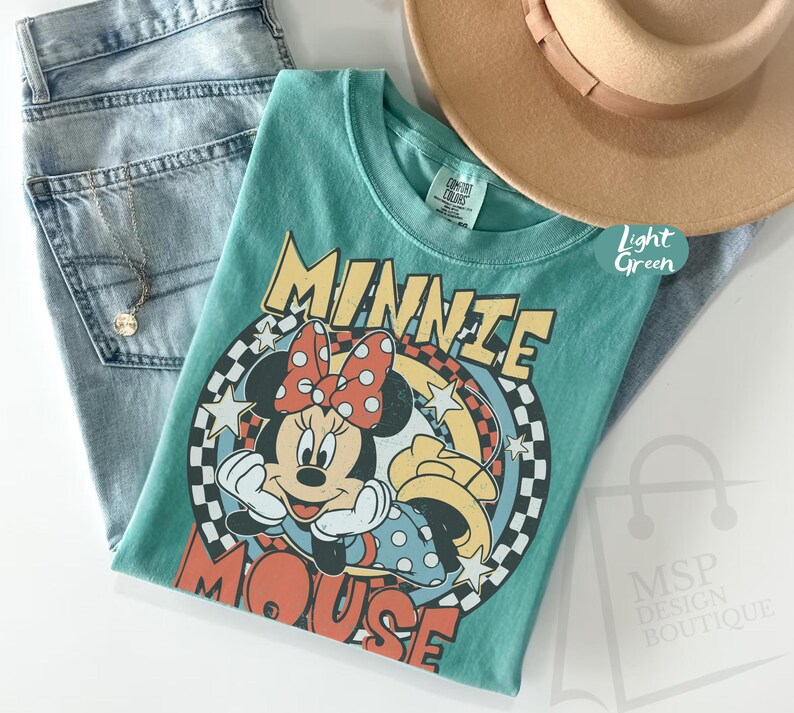 May include: A light green t-shirt featuring a graphic of Minnie Mouse with a red polka dot bow. The shirt is paired with light blue jeans and a tan fedora hat. A gold necklace is also visible.