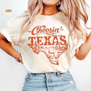 May include: Ivory-colored t-shirt with the text "Choosin' TEXAS" in a red, distressed font. The design features a map of Texas with cacti, a star, and a longhorn skull. The shirt is worn with blue denim shorts.