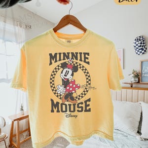 以下が含まれることがあります： ミニーマウスのグラフィックが特徴の黄色のボクシーTシャツ。デザインは、赤と白のドット柄のリボンとドレスを着たミニーマウスで、「MINNIE MOUSE」と「Disney」の文字が黒で描かれています。Tシャツは木製のハンガーに掛けられています。