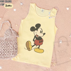 May include: A yellow tank top with a graphic of Mickey Mouse in red shorts and yellow shoes. The text "COMFORT COLORS TANK TOP Butter" is at the top left. A crocheted bag is on the left.