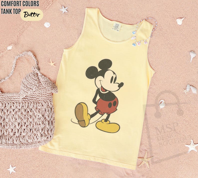 May include: A yellow tank top with a graphic of Mickey Mouse in red shorts and yellow shoes. The text "COMFORT COLORS TANK TOP Butter" is at the top left. A crocheted bag is on the left.