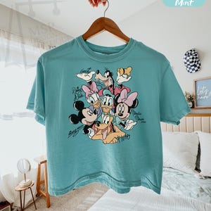 Puede incluir: Una camiseta turquesa Comfort Colors boxy con un gr&aacute;fico de Mickey Mouse, Minnie Mouse, Donald Duck, Daisy Duck, Goofy y Pluto. La camiseta tiene el texto "Mickey Mouse" y "Minnie Mouse" en cursiva. La camiseta est&aacute; colgada en una percha de madera.