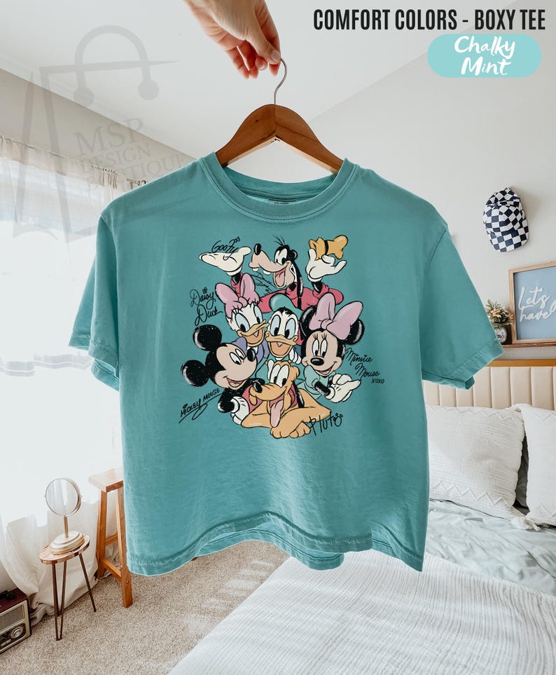 Puede incluir: Una camiseta turquesa Comfort Colors boxy con un gr&aacute;fico de Mickey Mouse, Minnie Mouse, Donald Duck, Daisy Duck, Goofy y Pluto. La camiseta tiene el texto "Mickey Mouse" y "Minnie Mouse" en cursiva. La camiseta est&aacute; colgada en una percha de madera.