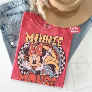 May include: A red t-shirt featuring a graphic of Minnie Mouse with a polka dot bow, stars, and the words "MINNIE MOUSE". The shirt is paired with light blue jeans and a tan hat.