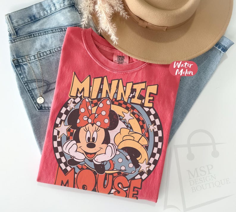 May include: A red t-shirt featuring a graphic of Minnie Mouse with a polka dot bow, stars, and the words "MINNIE MOUSE". The shirt is paired with light blue jeans and a tan hat.