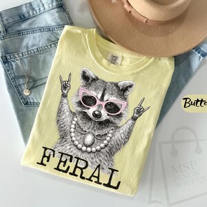 May include: A light yellow t-shirt with a graphic of a raccoon wearing pink sunglasses and a pearl necklace, with the word "FERAL" in black. The shirt is on top of blue jeans and near a tan hat.