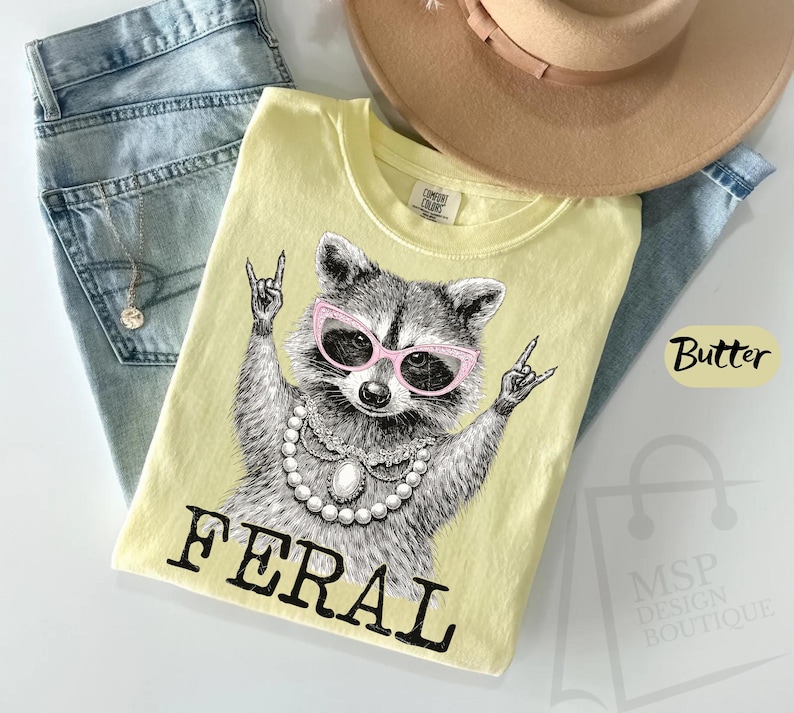 May include: A light yellow t-shirt with a graphic of a raccoon wearing pink sunglasses and a pearl necklace, with the word "FERAL" in black. The shirt is on top of blue jeans and near a tan hat.